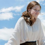 Georgie Henley is playing lucy. She is looking over the side of the Dawn Treader with a partially cloudy blue sky in the background.