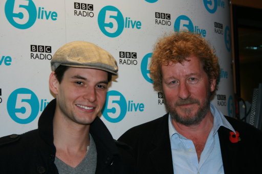 Ben Barnes stands on the left wearing a white hat while Sebastian Faulkes stands to the right. In the background is a white backdrop with the BBC Radio 5 Live logo tiled across the background.