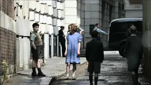 A candid, behind-the-scenes photo on a film set. Two child actors, one playing Digory and the other Polly, stand on a wet urban street. The boy wears a knickerbocker-style outfit with a sweater vest, and the girl wears a blue dress. Other cast and crew members are visible in the background.