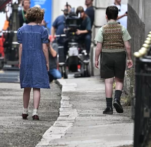 A behind-the-scenes photo of the two child actors, seen from behind, as they walk away from the camera down a narrow, wet alley on the film set. The boy is in knickerbocker-style shorts and a sweater vest, and the girl is in a blue dress with short sleeves.