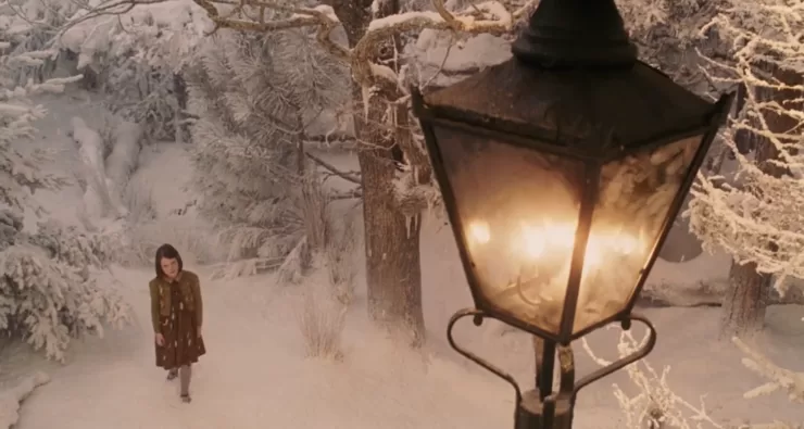 A screenshot from the 2005 Walden Media film, with a young girl standing in a snowy forest. An antique, gas-lit lamp-post with a warm, amber glow is prominent in the foreground. 
