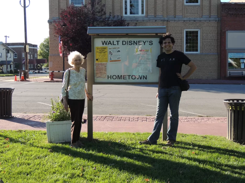 Janet Watkins and David Sutton stand on the grass by the sidewalk in Marceline, Missouri beside a sign reading Walt Disney's Hometown